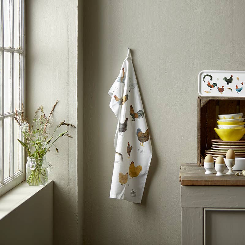 Ein Küchentuch von Koustrupco aus 100 % Bio-Baumwolle mit Hühnermotiven der Marke Koustrupco hängt an einer beigen Wand neben einem Fenster, in dem eine Vase mit Wildblumen steht. Daneben steht eine Holzkiste mit Geschirr, und vier Eier liegen in Eierbechern auf der grauen Arbeitsplatte – ein hervorragendes Beispiel für nachhaltiges Küchenzubehör.