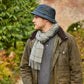 Ein Mann in einer olivgrünen Jacke und einem grauen Schal trägt den „Wachstropfen“-Krempenhut von Tweedmill Textiles – einen dunklen, gewachsten Baumwollhut mit breiter Krempe, hergestellt in Großbritannien –, während er im Freien steht und nach links blickt, im Hintergrund sind grünes Laub und eine Backsteinmauer zu sehen.