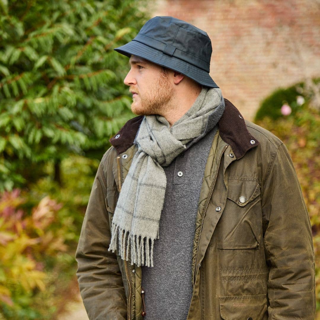 Ein Mann in einer olivgrünen Jacke und einem grauen Schal trägt den „Wachstropfen“-Krempenhut von Tweedmill Textiles – einen dunklen, gewachsten Baumwollhut mit breiter Krempe, hergestellt in Großbritannien –, während er im Freien steht und nach links blickt, im Hintergrund sind grünes Laub und eine Backsteinmauer zu sehen.