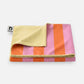 Das Notabag | Handtuch orange & rosa gestreift (78 × 160 cm) von Notabag ist hier gefaltet abgebildet und zeigt breite rosa und orangefarbene Streifen sowie eine zartgelbe Rückseite. Es besteht aus recyceltem Polyester und ist an einer Ecke mit einem weißen Etikett mit schwarzem Logo versehen.
