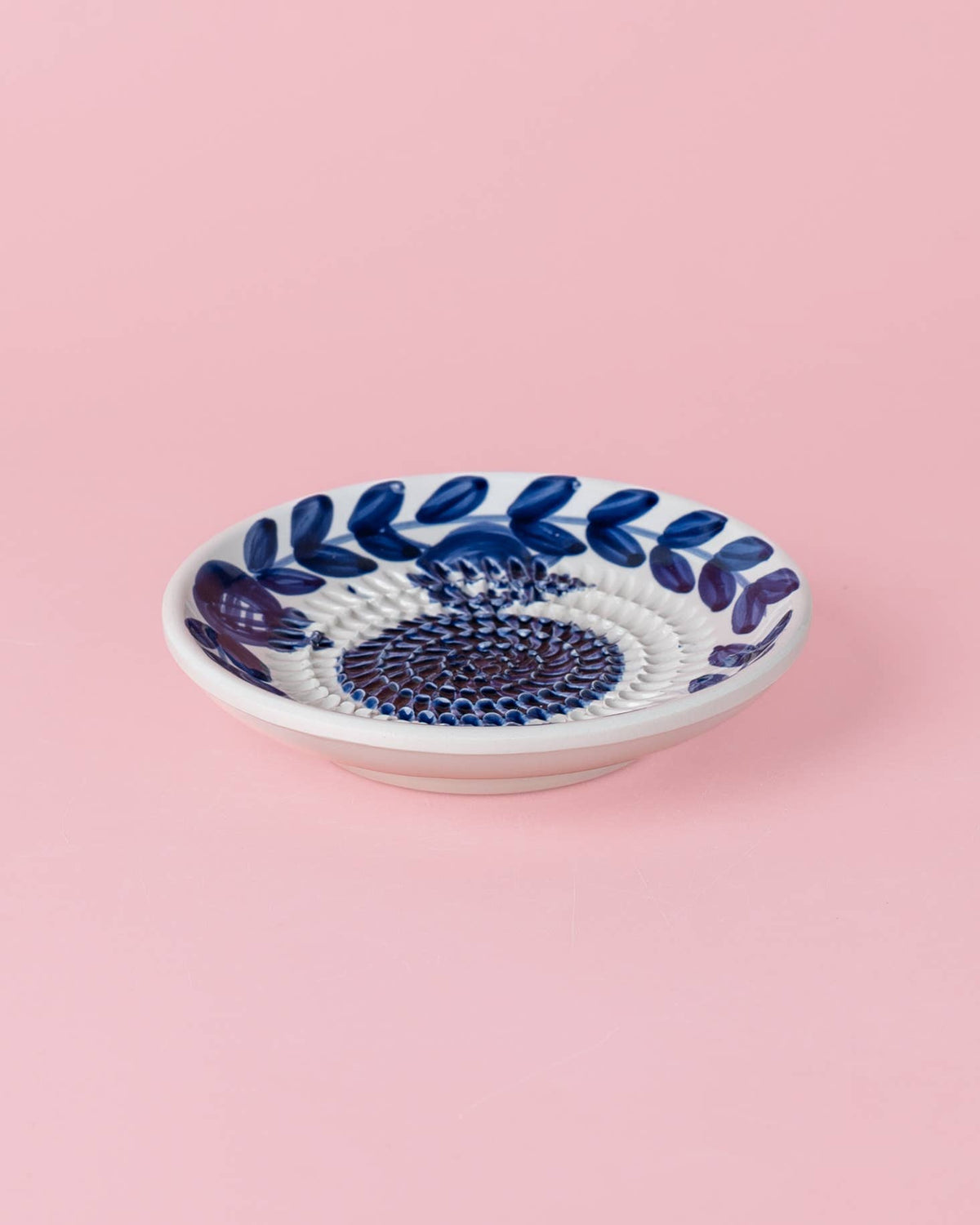 Eine Alfar Tierra Cocida | Kleine Keramik-Knoblauchreibe „Nerja“, traditionell von Hand in Blau bemalt, ruht auf einem schlichten hellrosa Hintergrund.