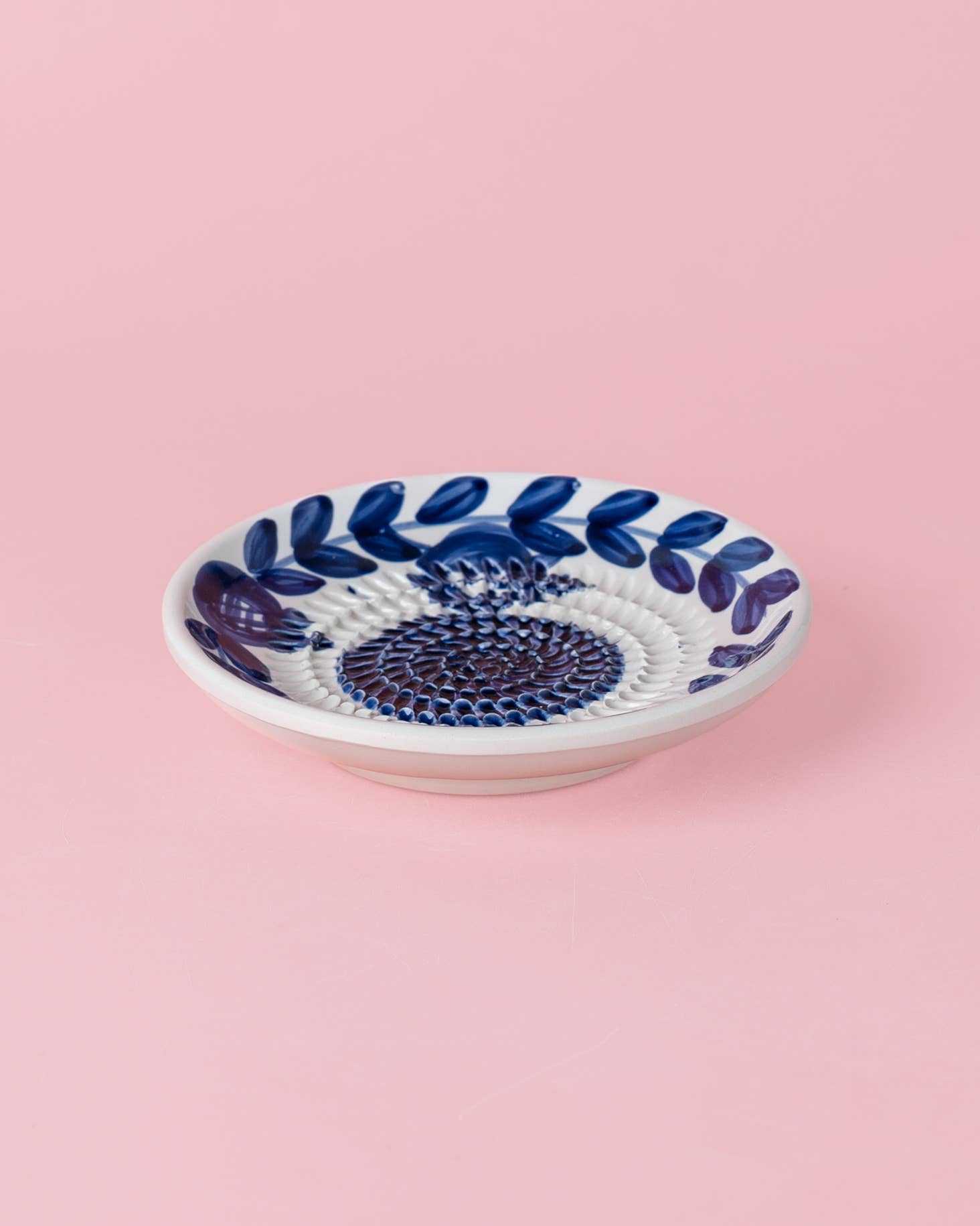 Eine Alfar Tierra Cocida | Kleine Keramik-Knoblauchreibe „Nerja“, traditionell von Hand in Blau bemalt, ruht auf einem schlichten hellrosa Hintergrund.