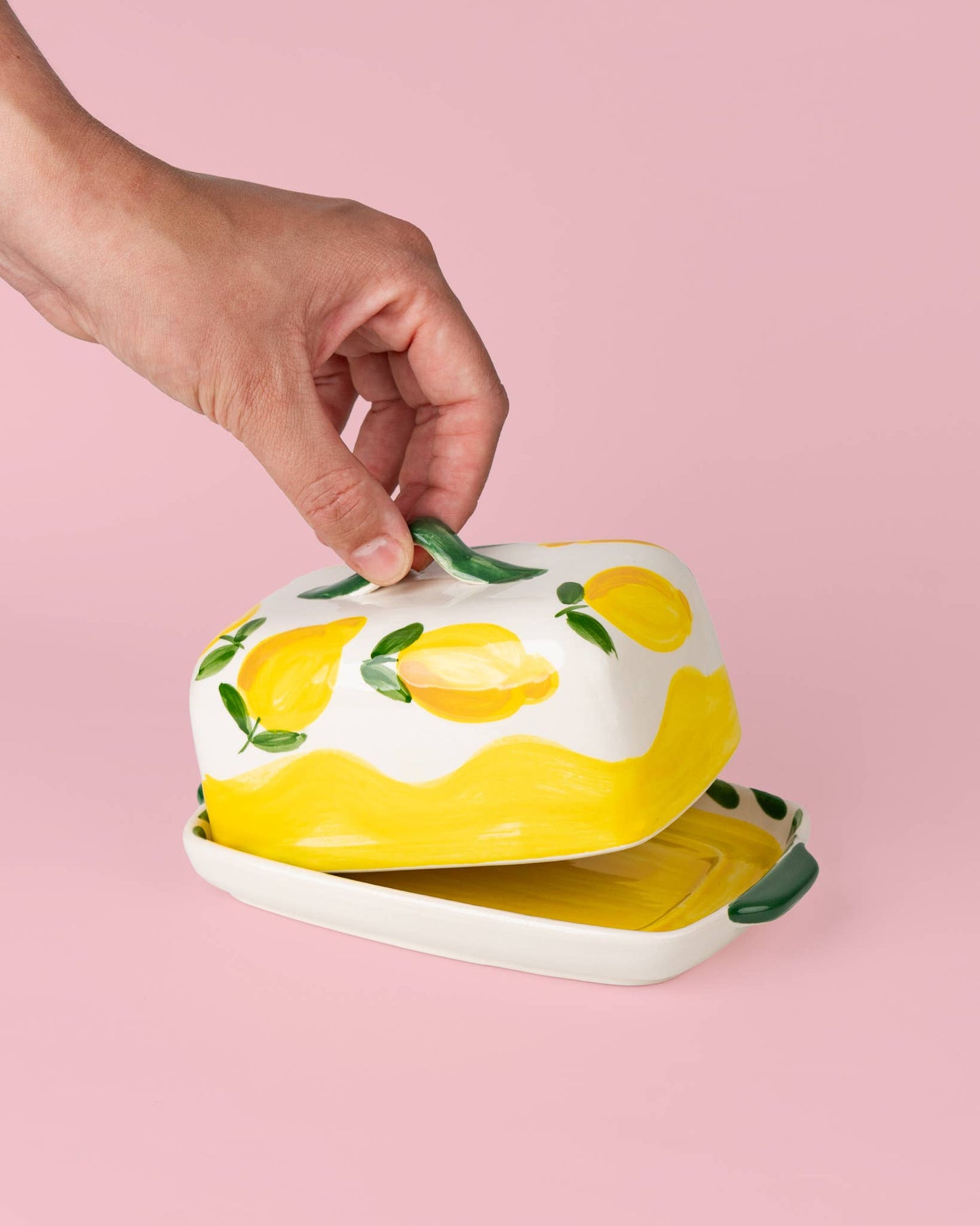 Eine Hand hebt den Deckel der Alfar Tierra Cocida | Handgefertigte Keramik-Butterdose „Limone“ in Gelb und Weiß an, deren Oberfläche mit handgemalten Zitronenmotiven und grünen Blättern verziert ist und den Charme der mediterranen Alfar Tierra Cocida-Tischwaren vor einem zartrosa Hintergrund zum Leben erweckt.