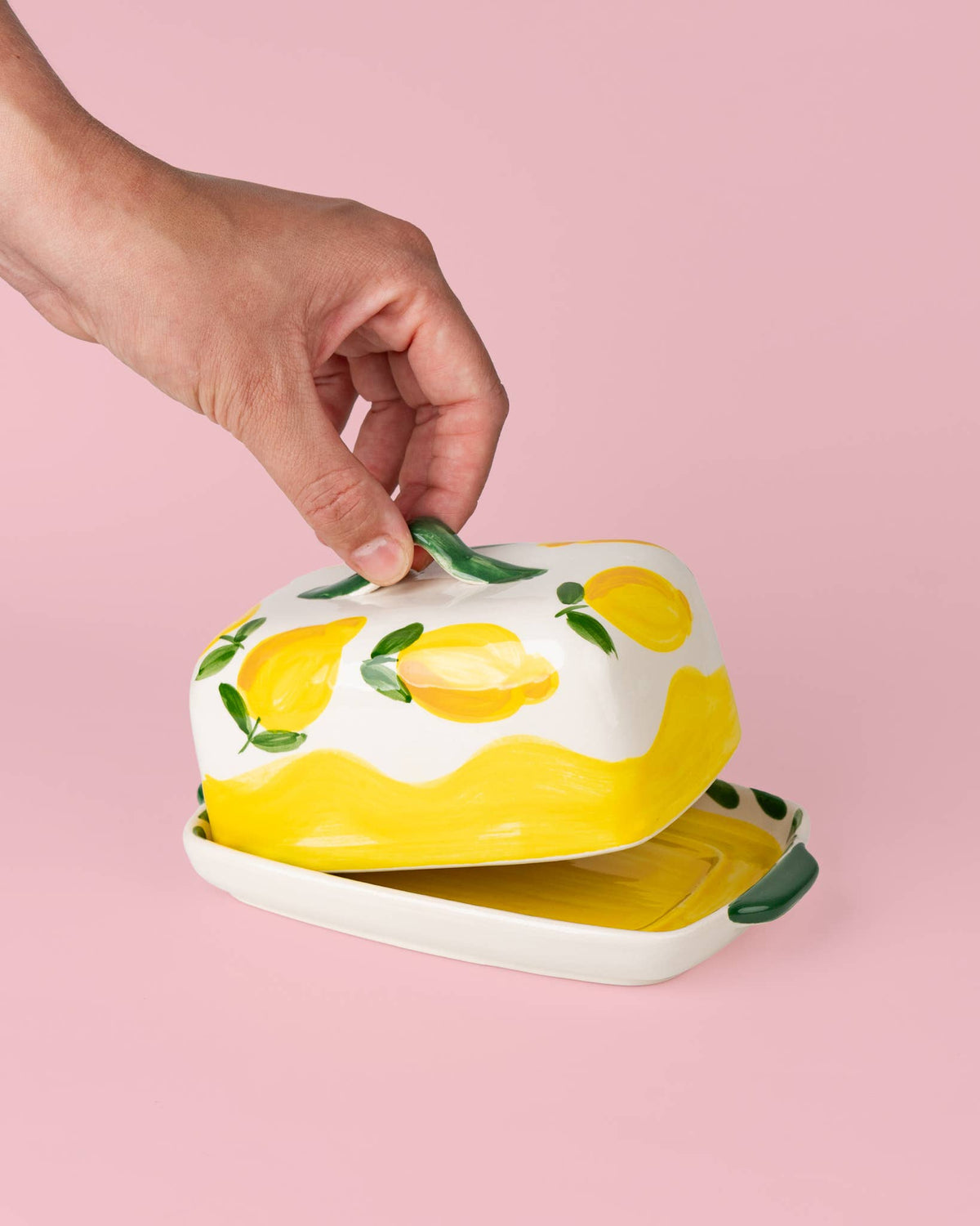 Eine Hand hebt den Deckel der Alfar Tierra Cocida | Handgefertigte Keramik-Butterdose „Limone“ in Gelb und Weiß an, deren Oberfläche mit handgemalten Zitronenmotiven und grünen Blättern verziert ist und den Charme der mediterranen Alfar Tierra Cocida-Tischwaren vor einem zartrosa Hintergrund zum Leben erweckt.