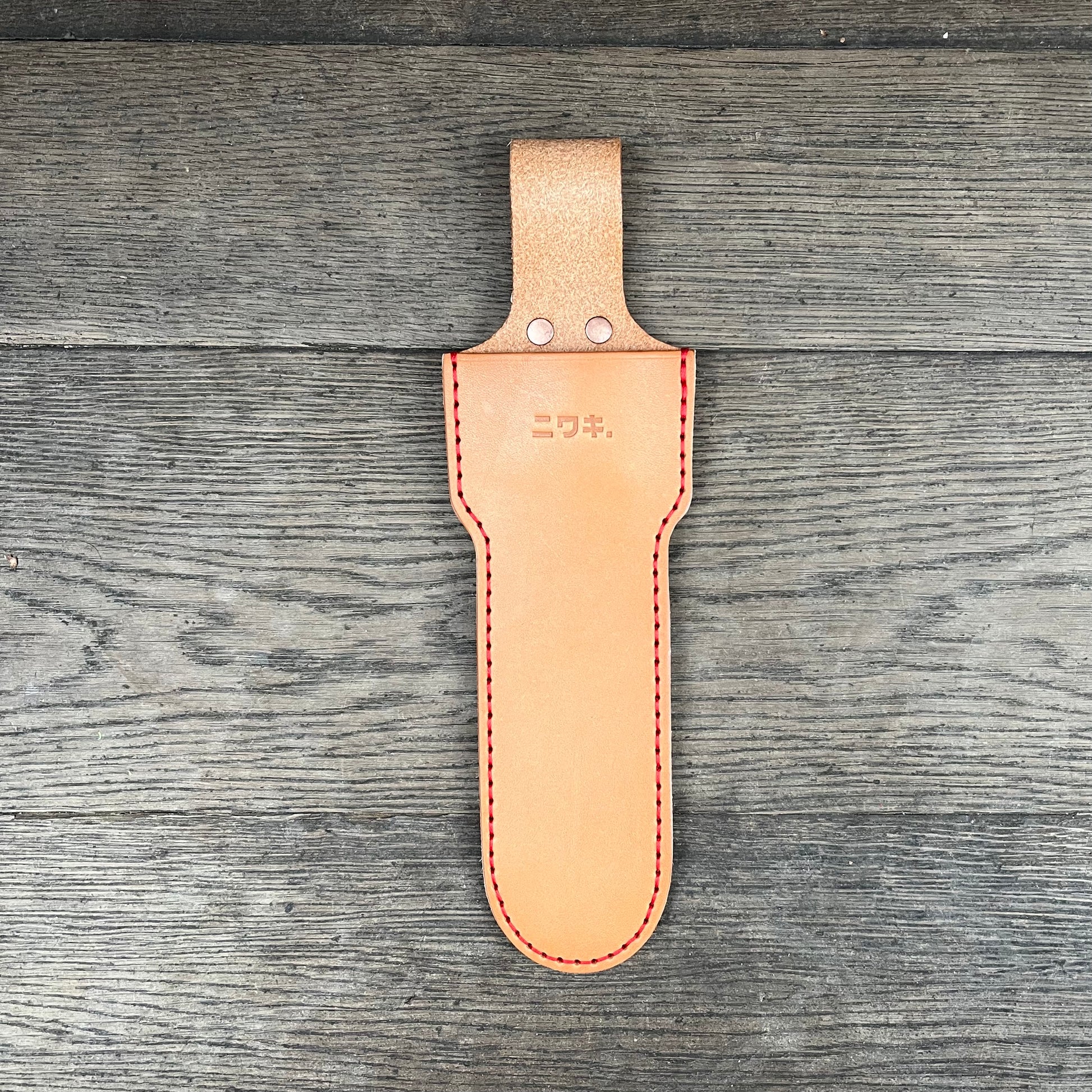 Die Lederschutzhülle für große Scheren Niwaki | 'Long Holster' aus hellbraunem Leder mit roten Nähten und Metallnieten ruht auf Holz und ist mit einer japanischen Textprägung im oberen Bereich und einer Gürtelschlaufe versehen. Marke: Niwaki.