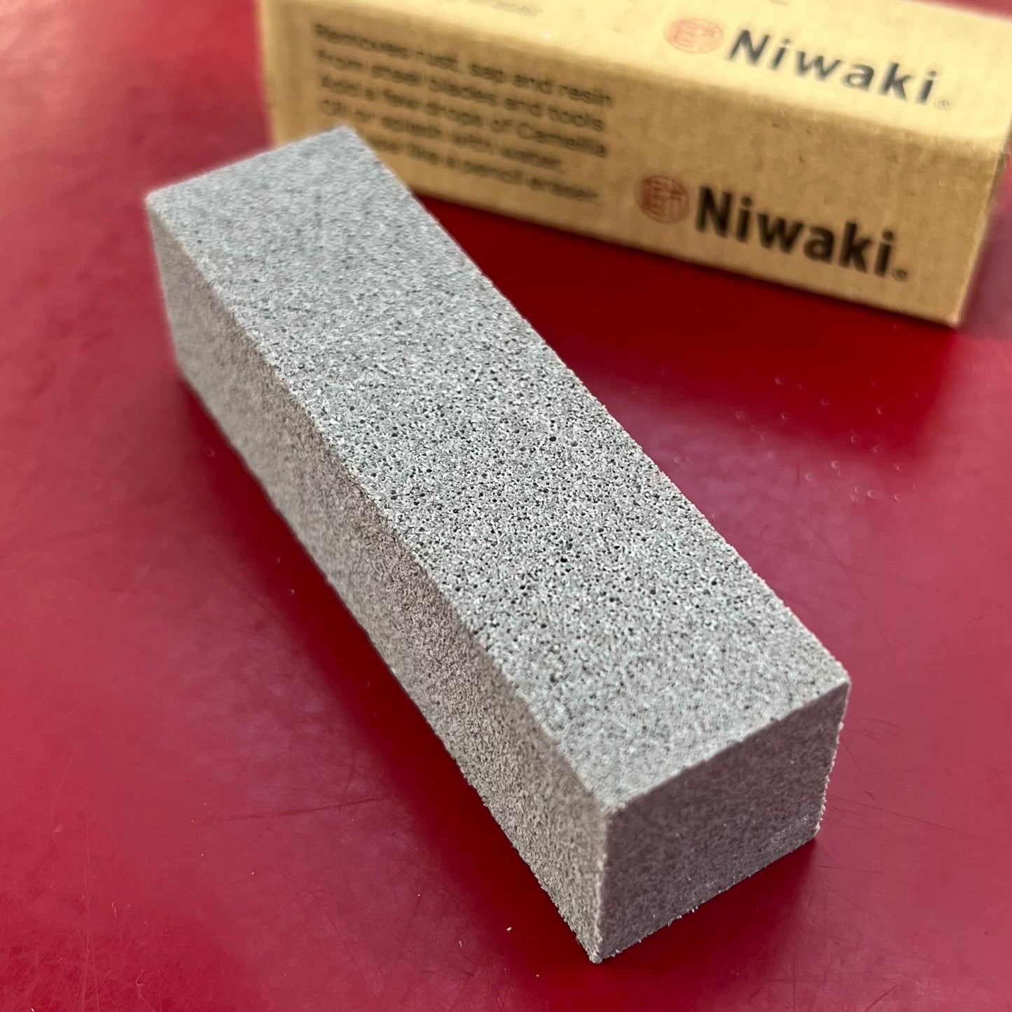 Ein grauer Niwaki 'Mini Clean Mate' Rost- und Harzentferner steht auf einer roten Fläche, dahinter die Pappschachtel - ideal zum Reinigen von Gartenwerkzeugen oder zum Entfernen von Rost und Harz von Kohlenstoffstahlwerkzeugen.