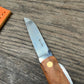 Das Niwaki | 'Kotoh X Niwaki Knife' Taschenmesser mit Schutzhülle aus Leder von Niwaki, ausgestattet mit einem Holzgriff und Edelstahlklinge, liegt auf verwittertem Holz neben einem orangenen Lederetui - ideal für Gartenarbeit oder den Alltag.