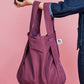 Eine Person, die eine dunkle Jeansjacke trägt, hält die Notabag | Wandelbare Tasche und Rucksack „Pflaume“ von Notabag – eine wiederverwendbare Tasche mit violettem Karomuster und einem kleinen weißen Logo-Etikett – vor einem rosa Hintergrund.