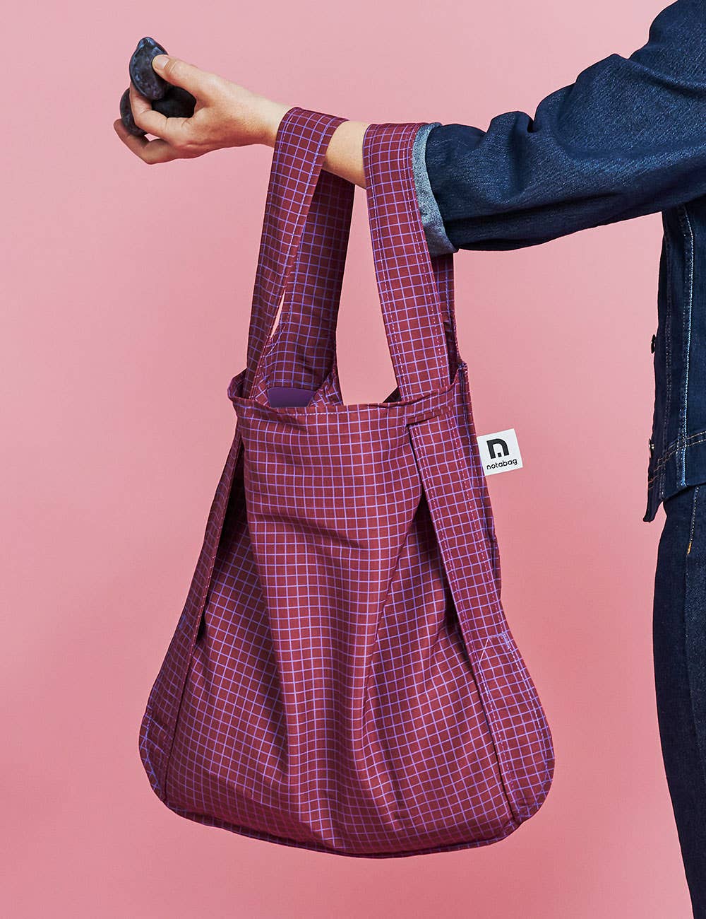 Eine Person, die eine dunkle Jeansjacke trägt, hält die Notabag | Wandelbare Tasche und Rucksack „Pflaume“ von Notabag – eine wiederverwendbare Tasche mit violettem Karomuster und einem kleinen weißen Logo-Etikett – vor einem rosa Hintergrund.