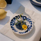 Die Alfar Tierra Cocida | Kleine Keramik-Knoblauchreibe „Nerja“, handbemalt mit traditionellen blau-weißen Blumenmotiven, ist abgebildet, wie sie eine gelbe Blume und frische Kräuter auf einer beigen Serviette hält. Daneben liegen Zitronen und eine weitere Schale auf einer weißen Tischdecke, alles im hellen Sonnenlicht.