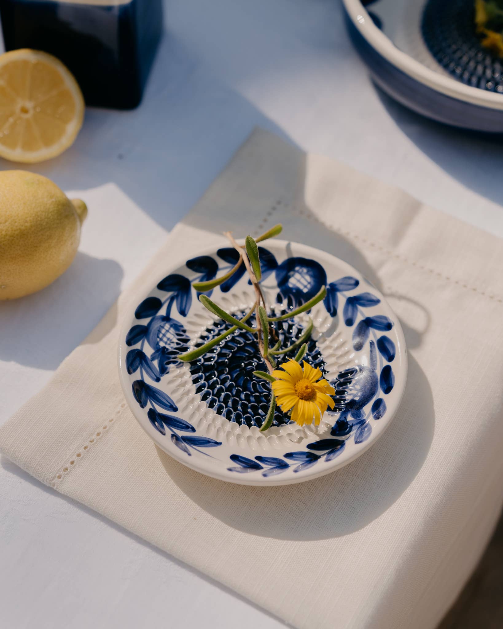 Die Alfar Tierra Cocida | Kleine Keramik-Knoblauchreibe „Nerja“, handbemalt mit traditionellen blau-weißen Blumenmotiven, ist abgebildet, wie sie eine gelbe Blume und frische Kräuter auf einer beigen Serviette hält. Daneben liegen Zitronen und eine weitere Schale auf einer weißen Tischdecke, alles im hellen Sonnenlicht.