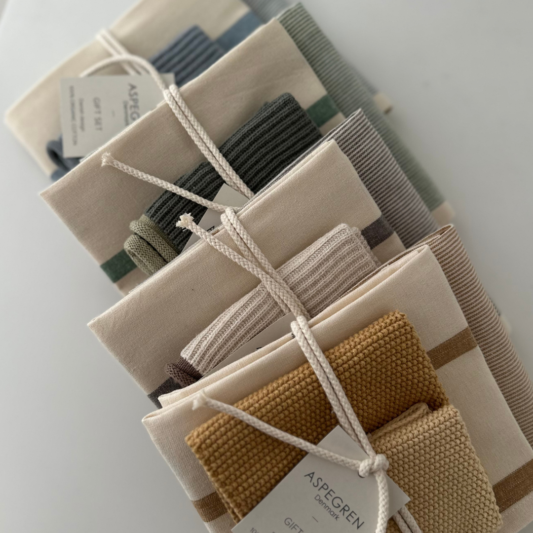 Vier Sets von Aspegren Denmark | „Grünes Küchentuch“ 3er-Geschenkset, bei denen jeweils ein Bündel gefalteter Küchentücher mit einer Schnur zusammengebunden und mit „Aspegren Denmark“ beschriftet ist, sind diagonal auf einer hellen Unterlage angeordnet. Die Tücher sind in neutralen und erdigen Farben gehalten und weisen gestreifte sowie strukturierte Muster auf.