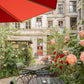 Ein Straßencafé mit schwarzen Metalltischen und -stühlen auf Kies, blühenden Rosen und Grünpflanzen ist in "Stadtoasen in Berlin | Die schönsten Gartencafés, Biergärten und Draußenrestaurants" vom Bebra Verlag abgebildet. Ein roter Regenschirm beschattet die Gäste, dahinter befindet sich ein altes Gebäude mit Fenstern.