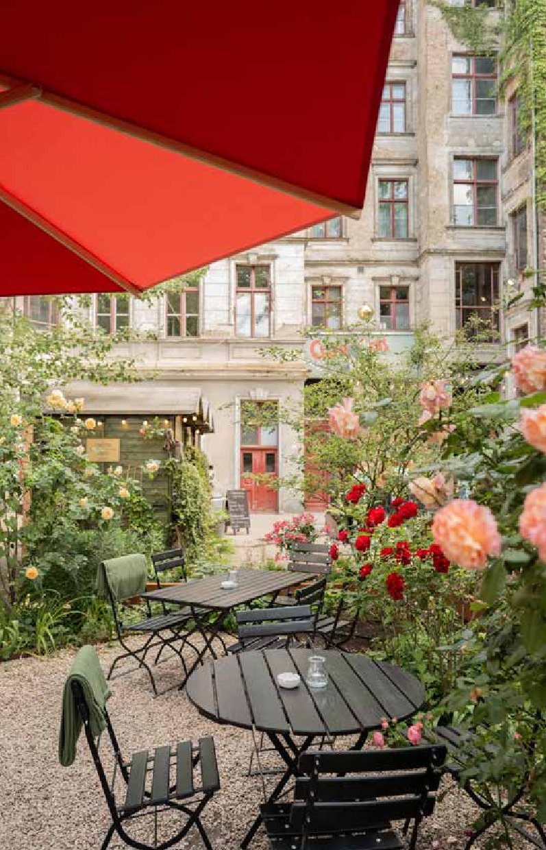 Ein Straßencafé mit schwarzen Metalltischen und -stühlen auf Kies, blühenden Rosen und Grünpflanzen ist in "Stadtoasen in Berlin | Die schönsten Gartencafés, Biergärten und Draußenrestaurants" vom Bebra Verlag abgebildet. Ein roter Regenschirm beschattet die Gäste, dahinter befindet sich ein altes Gebäude mit Fenstern.