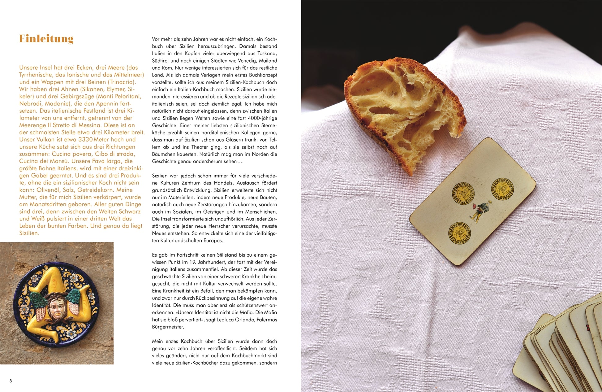 Ein Laib Brot und eine Tarotkarte liegen auf einem weißen Tuch neben einem Teller mit Oliven, Gemüse und Zitronenscheiben und erinnern an sizilianische Küche. Das aufgeschlagene Buch "Sizilien in meiner Küche | Eine kulinarische Rundreise" aus dem DK Verlag zeigt den deutschen Text.