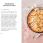 Ein runder Apfelkuchen in einer mit Pergament ausgelegten Form steht auf einer beigen Fläche neben einer Seite aus dem DK Verlag "Kleine Kuchen | 50 Rezepte für Mini-Kuchen, kleine Torten & mehr" mit veganen und glutenfreien Rezepten und Zutaten für Apfelkuchen aus der Toskana.