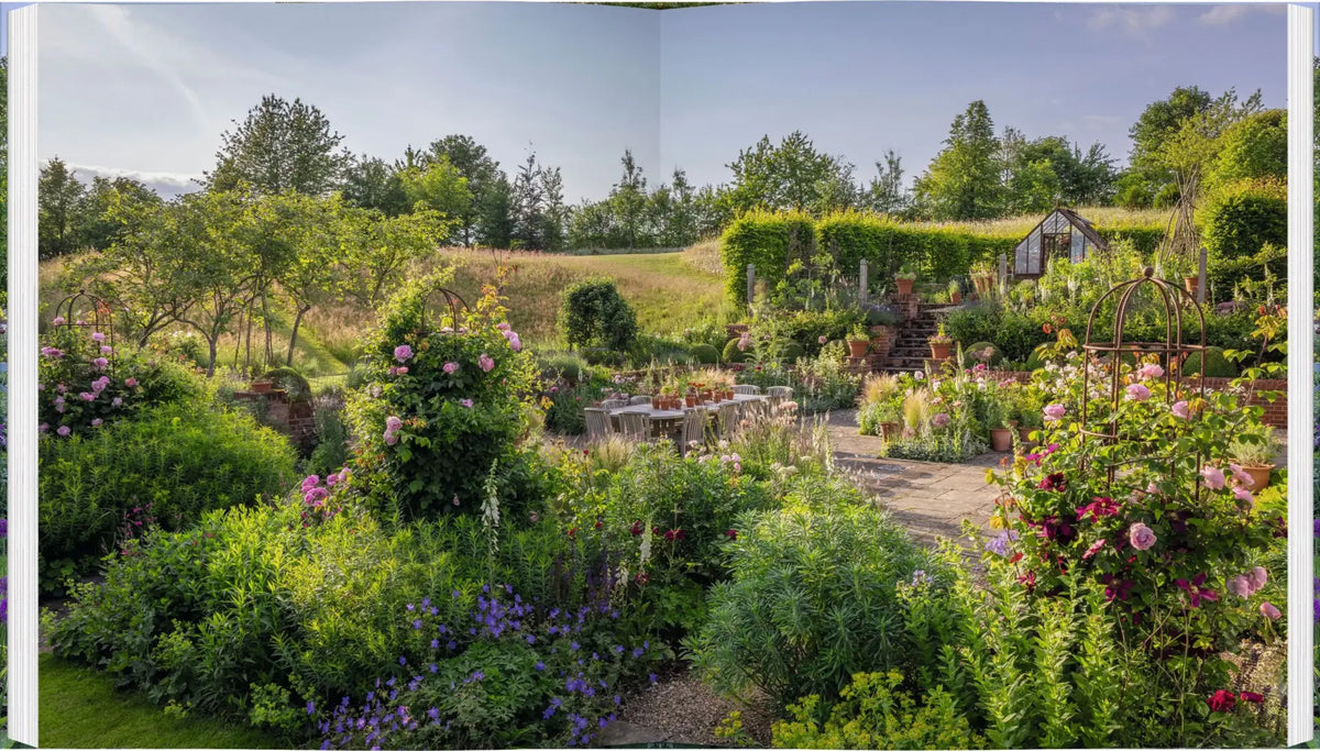 Ein sonnendurchfluteter englischer Cottage-Garten mit blühenden Blumenbeeten, einer Steinterrasse und einem Gewächshaus am Hang aus dem Buch Die schönsten Cottage-Gärten Englands, das klassische englische Gartengestaltung präsentiert.