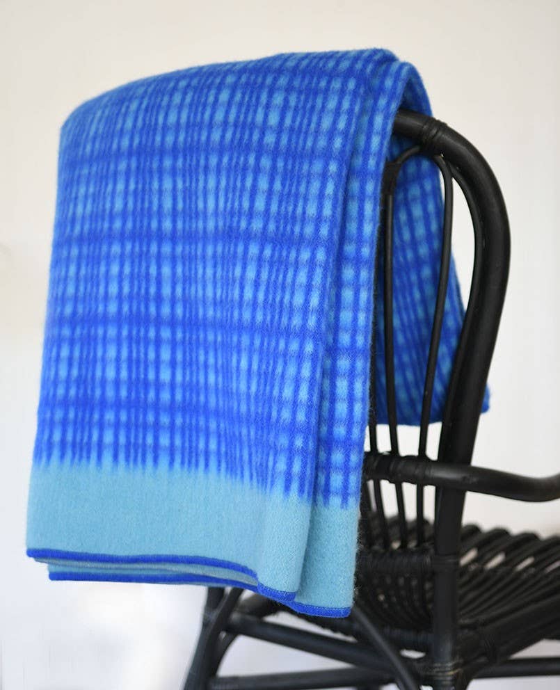 Die Sofie Sjöström 'Darling' blaue Tagesdecke aus Lammwolle von Sofie Sjöström Design ist über einen schwarzen, geflochtenen Stuhl drapiert und steht vor einem schlichten weißen Hintergrund.