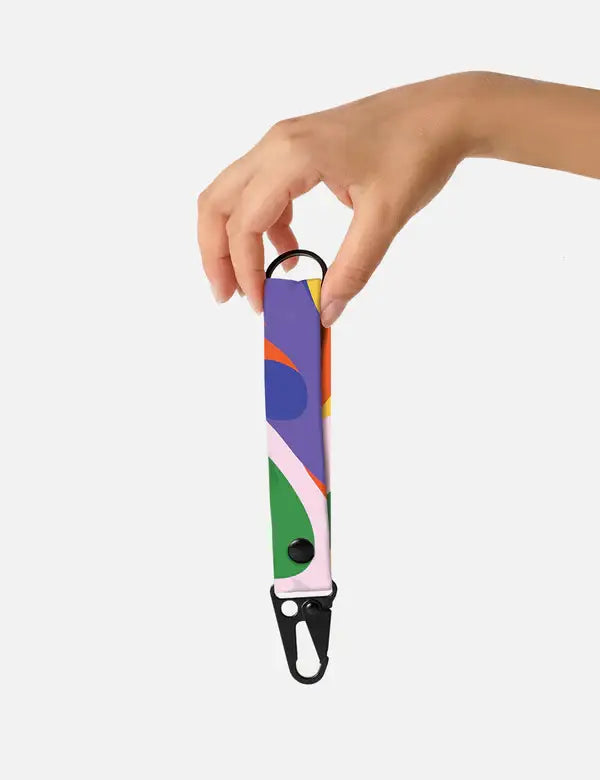 Eine Hand hält den „Notabag – Schlüsselanhänger – Flow“ von Notabag, der sich durch ein farbenfrohes, abstraktes Muster und einen schwarzen Metallclip auszeichnet und vor einem schlichten weißen Hintergrund als nachhaltiges Accessoire präsentiert wird.