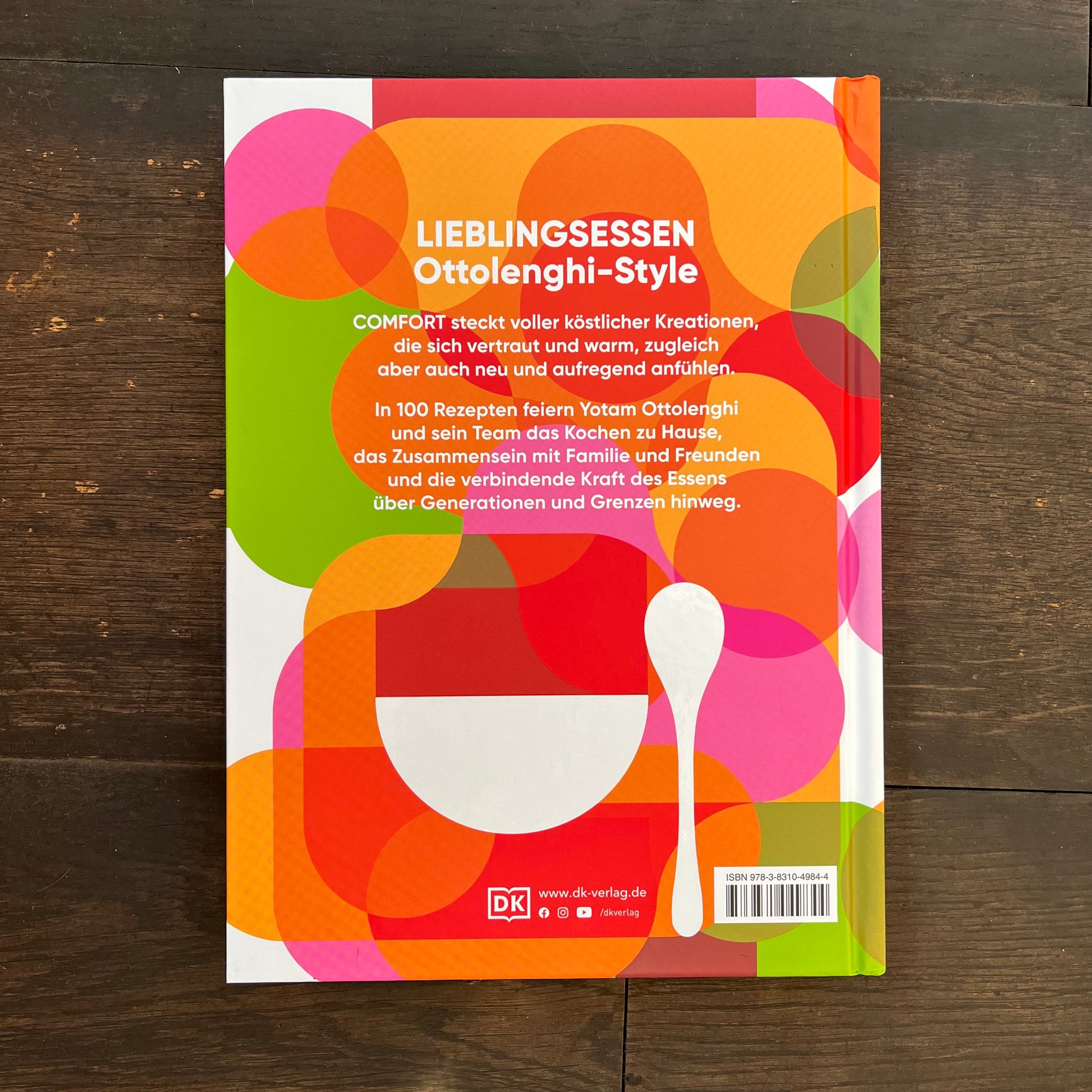 Die Rückseite des DK Kochbuch Ottolenghi "Comfort" zeigt lebendige, sich überlappende Kreise, deutschen Text, eine weiße Löffelgrafik und Verlagsangaben. Es zeigt Genuss und natürliche Zutaten auf einem dunklen Holzhintergrund.