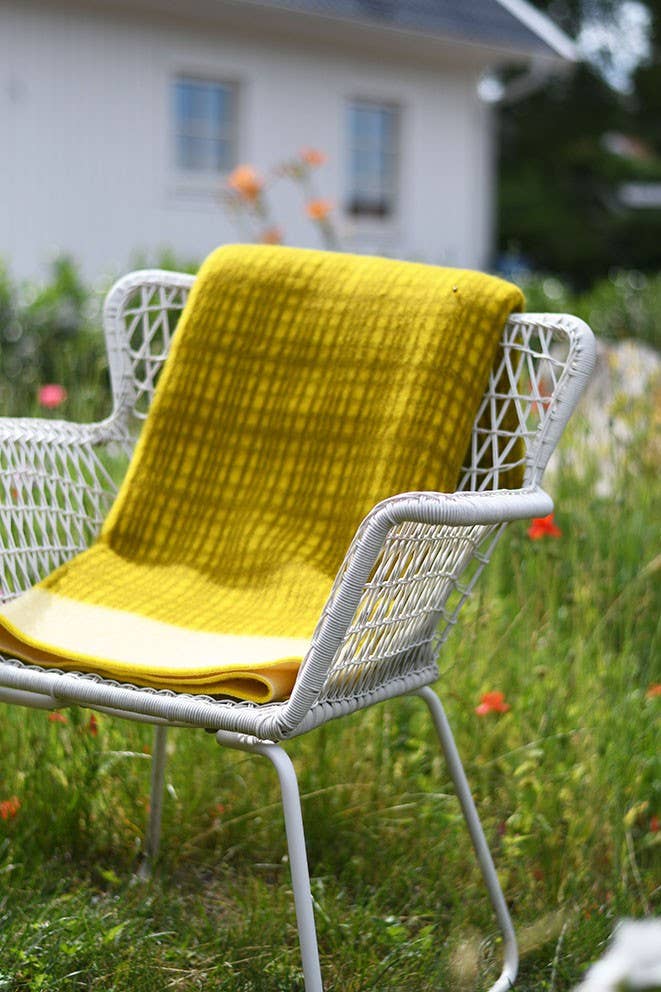 Ein weißer Metallstuhl mit geflochtenem Muster sitzt im Gras, drapiert mit der Sofie Sjöström Design 'Darling' gelben Tagesdecke aus Lammwolle. Im Hintergrund sind verschwommene Blumen und ein weißes Haus zu sehen.