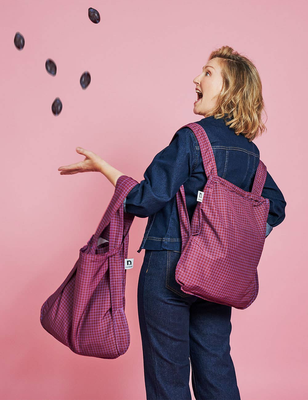 Eine Frau in einem Jeansoutfit lächelt, während sie kleine dunkle Gegenstände in die Luft wirft. Sie trägt zwei „Notabag | Wandelbare Tasche und Rucksack ‚Pflaume‘“-Taschen von Notabag über der Schulter und steht vor einem rosa Hintergrund.