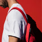 Ein bärtiger Mann in einem weißen T-Shirt trägt die Notabag | „Original Red“ – Tasche und Rucksack in einem von Notabag – über einer Schulter und steht vor einem leuchtend roten Hintergrund, wobei sein Gesicht teilweise außerhalb des Bildausschnitts liegt.