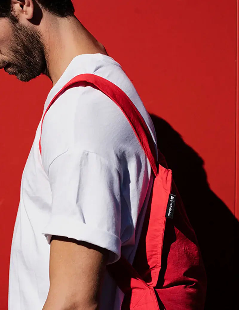 Ein bärtiger Mann in einem weißen T-Shirt trägt die Notabag | „Original Red“ – Tasche und Rucksack in einem von Notabag – über einer Schulter und steht vor einem leuchtend roten Hintergrund, wobei sein Gesicht teilweise außerhalb des Bildausschnitts liegt.