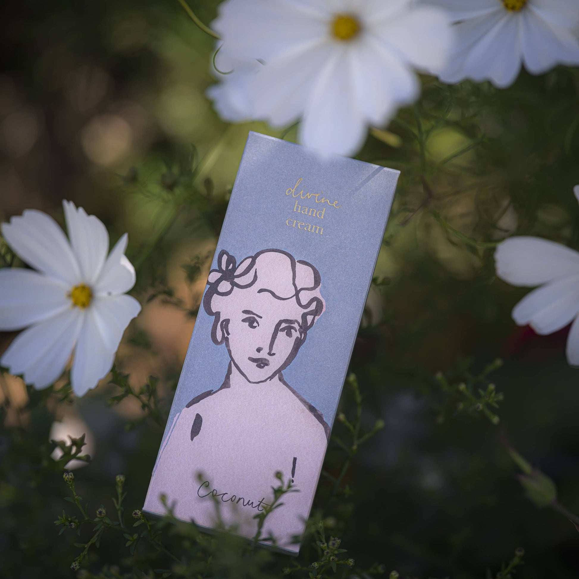 Eine Schachtel der "Kokosnuss"-Handcreme von Archivist Gallery, auf deren Verpackung ein Frauengesicht abgebildet ist, ist von weißen Blumen und Grünzeug umgeben.