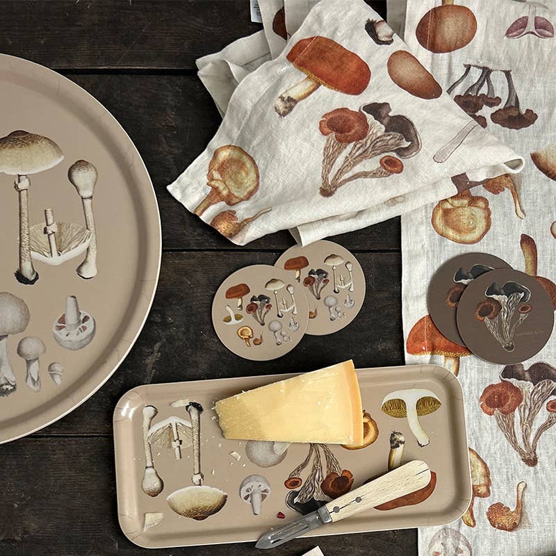 Auf einer dunklen Holzfläche sind verschiedene beigefarbene Tabletts, Untersetzer und Tischwäsche mit bezaubernden Pilzmotiven – Pilzillustrationen – angeordnet. Ein Stück Käse und ein Messer liegen auf einem Koustrupco-Serviertablett 32 x 15 cm „Pilz“ von Koustrupco, das meisterhaft aus elegantem Birkenfurnier gefertigt wurde.