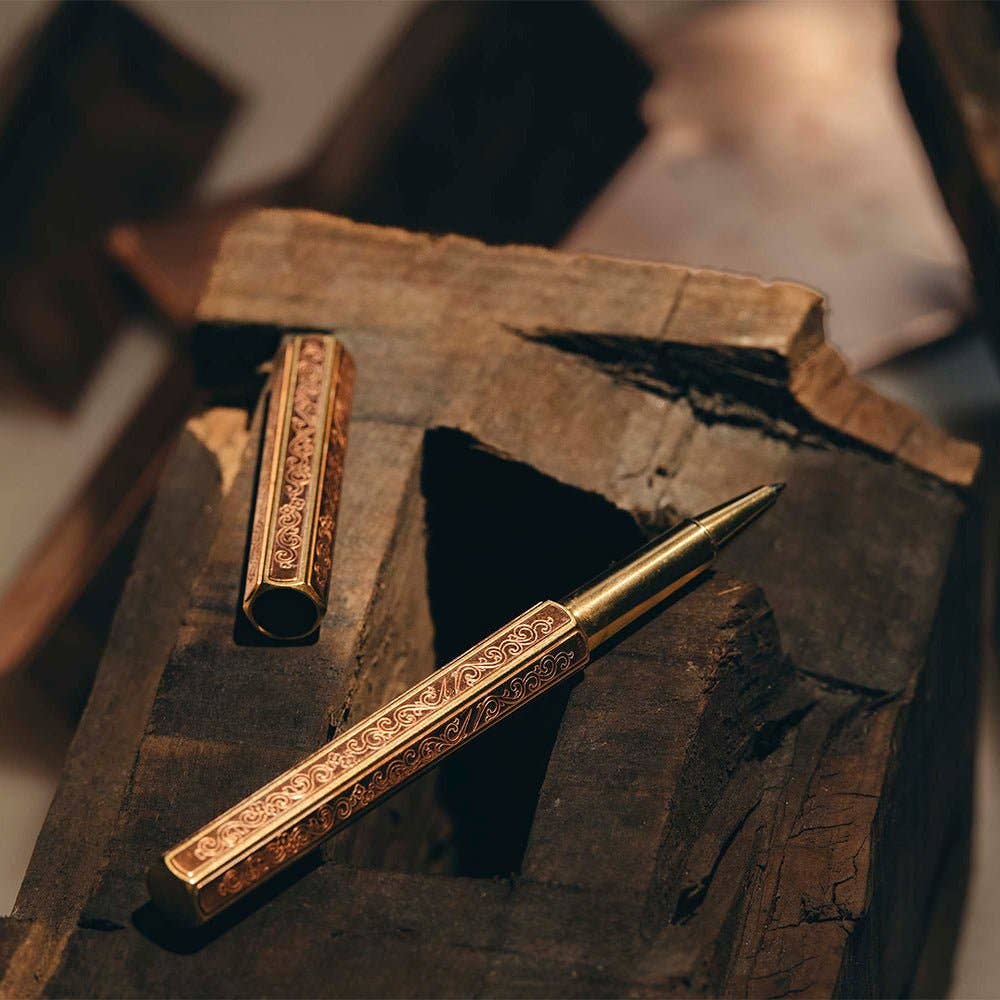 Der ENTREPÔT YSTUDIO | Klassische Renaissance KAZARI KANAGU ORCHID Tintenroller mit verschnörkelten, goldenen Details und Metallgravurkunst ruht auf dunklem Holz unter warmem Licht, sein Deckel daneben hebt filigrane Gravuren hervor.