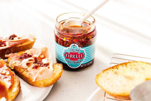 Ein Glas Firelli | Chili Crunch 200g Glas mit einem Löffel steht neben getoastetem Brot und mit Prosciutto und knusprigen Chiliflocken belegten Scheiben, alles auf einer weißen Fläche angerichtet.