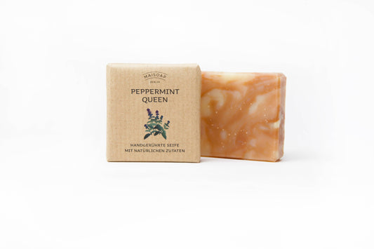 Ein Stück Maisoap | „Peppermint Queen“ Seife mit roter Tonerde von Maisoap Berlin und eine beschriftete Schachtel mit einer Illustration von Pfefferminze vor weißem Hintergrund. Auf der Verpackung befinden sich Texte in Englisch und Deutsch, die dieses Produkt als eines unserer natürlichen Pflegeprodukte hervorheben.