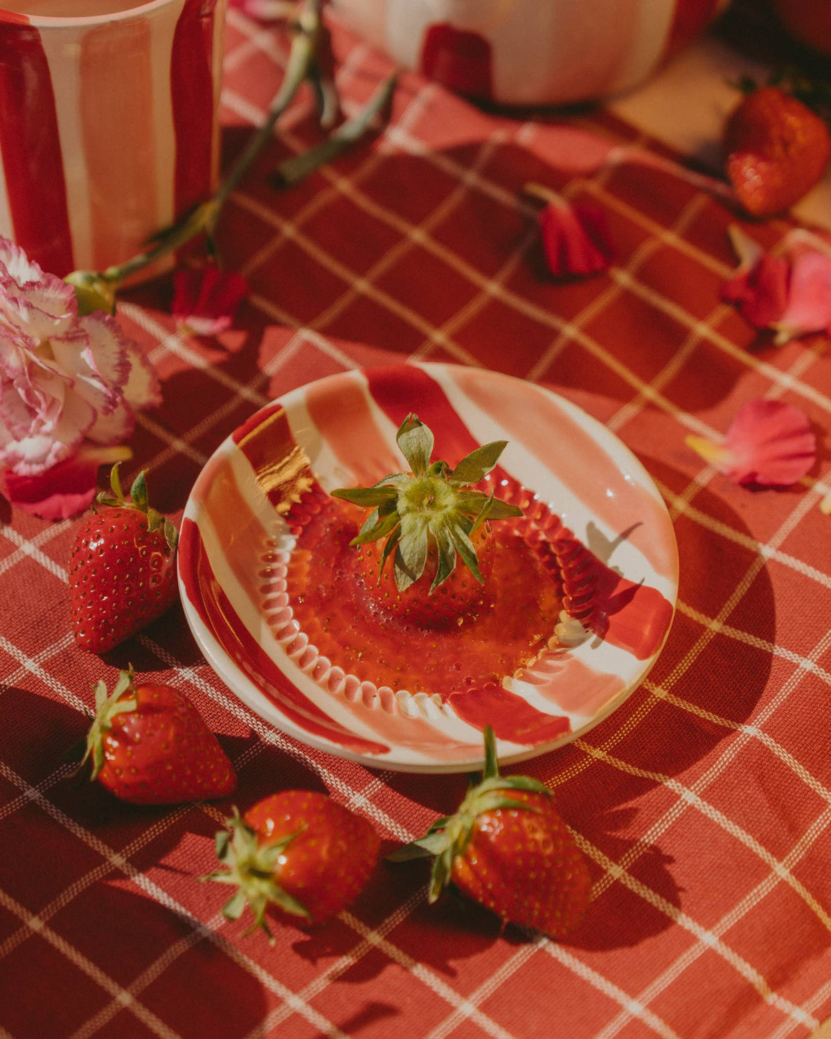 Ein Teller mit dem Motiv „Alfar Tierra Cocida | Kleine keramische Knoblauchreibe in Rosa & Rot“ von Alfar Tierra Cocida, verziert mit einer Erdbeere in der Mitte und umgeben von weiteren Erdbeeren und rosa Blütenblättern auf einer rot karierten Tischdecke im warmen Sonnenlicht, unterstreicht die Schönheit handgefertigter Töpferkunst als charmantes Küchenaccessoire.