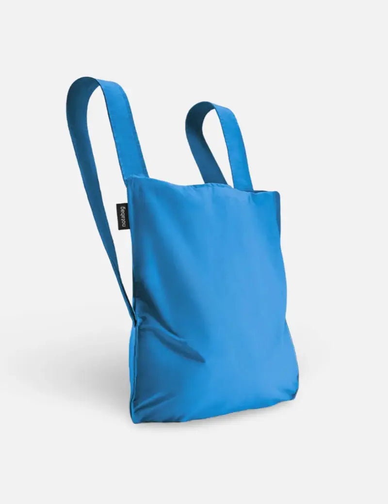 Die Notabag | „Original Blue“-Tasche von Notabag ist eine vielseitige blaue Tragetasche, ein Rucksack und eine Handtasche in einem – mit zwei breiten Tragegriffen, einem minimalistischen Design und einem kleinen schwarzen Etikett an der Seite – perfekt für stilvolle und nachhaltige Transportbedürfnisse.