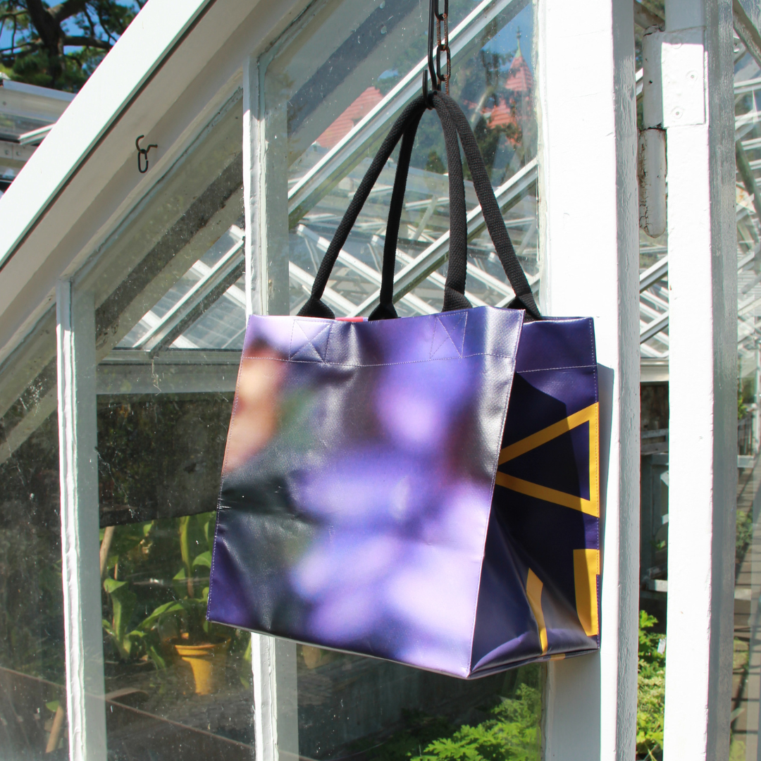 Die Upcycling Tasche 'Aster' aus Werbeplane von Königliche Gartenakademie mit schwarzen Griffen hängt im sonnenbeschienenen Gewächshaus, umgeben von üppigen Pflanzen und Glasscheiben.