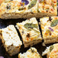Goldene, fluffige Focaccia mit Rosmarin, Salbei und essbaren Blüten wird wunderschön auf Holz serviert - ideal für Deko-Ideen Frühling oder Osterfest. Entdecken Sie mehr in "Frühlingstraum | Rezepte für Frühlingsgefühle und österliche Anlässe" vom Callwey Verlag.