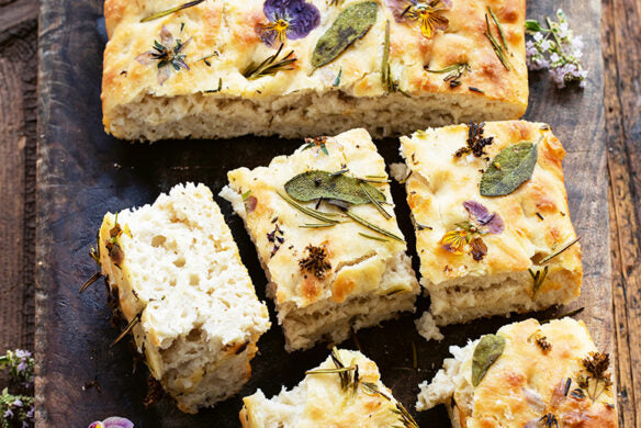 Goldene, fluffige Focaccia mit Rosmarin, Salbei und essbaren Blüten wird wunderschön auf Holz serviert - ideal für Deko-Ideen Frühling oder Osterfest. Entdecken Sie mehr in "Frühlingstraum | Rezepte für Frühlingsgefühle und österliche Anlässe" vom Callwey Verlag.