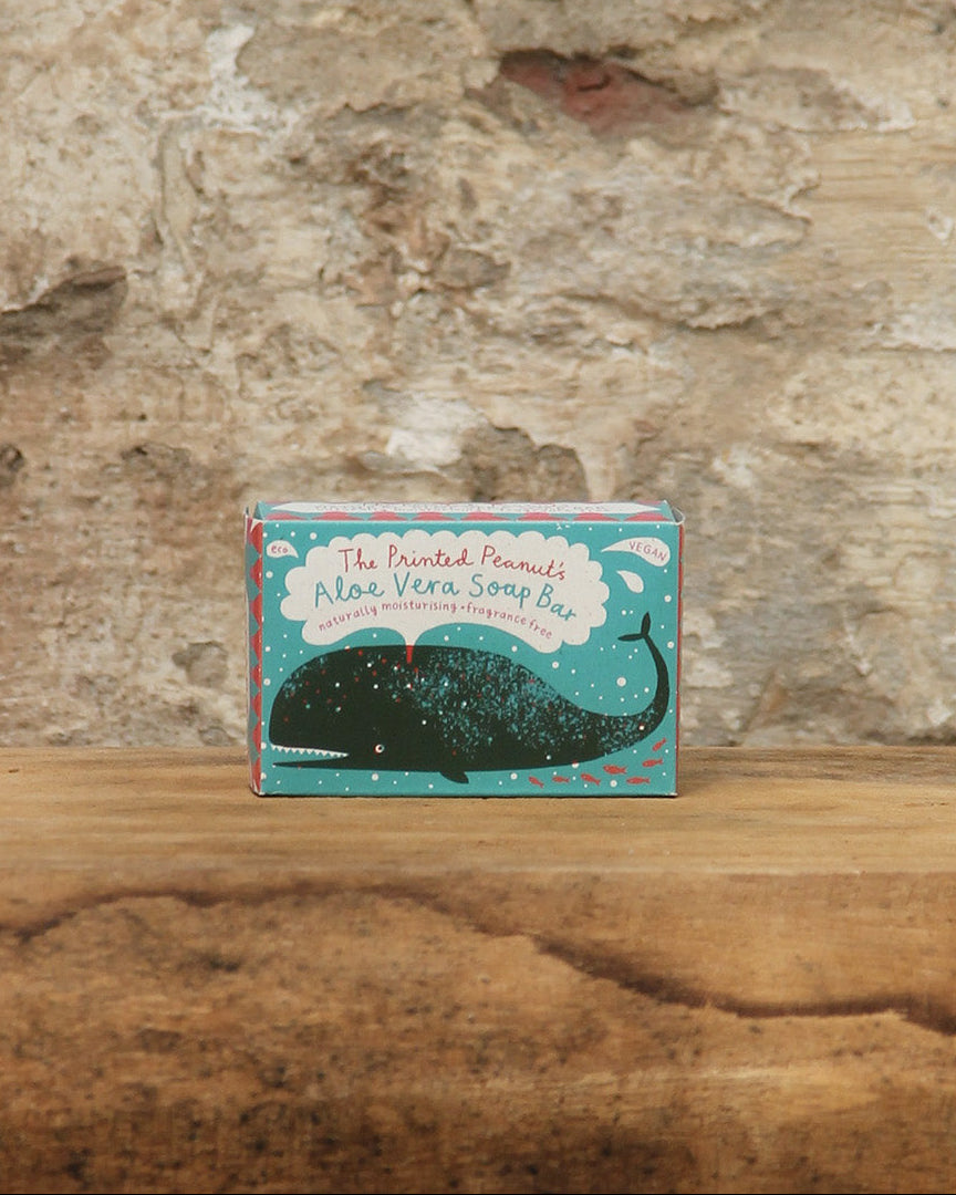 Eine Schachtel mit The Printed Peanut | Aloe Vera Seife von The Printed Peanut ruht auf einer hölzernen Oberfläche vor einer verwitterten Steinmauer. Die Verpackung zeigt einen dunklen Wal auf Petrol mit weißen und roten Akzenten.