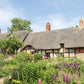 Inspiriert von "Gärtnern wie Gott in England" aus dem Callwey Verlag, stellen Sie sich ein traditionelles englisches Cottage mit Strohdach, sichtbarem Fachwerk und einem üppigen Garten mit grünen Pflanzen und violetten Blumen unter blauem Himmel vor.