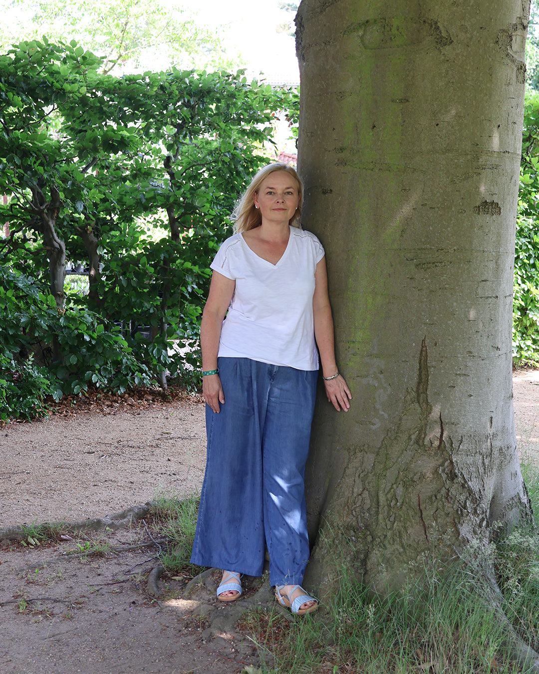 Eine Frau mit langen blonden Haaren, gekleidet in ein weißes Hemd, eine blaue Hose mit weitem Bein und Sandalen, steht an einem Baum - bereit für den Fr. 27.02.2026 | 09.30-12.30 HALBTAGESKURS Grundkurs "Grüner Daumen" der Königlichen Gartenakademie.