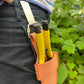 Eine Person in dunkler Hose befestigt die Niwaki 'Belt Clip' Praktische Halterung für Niwaki Single Holster von Niwaki an ihrer Tasche, während sie ein Gartengerät mit gelbem Griff und grünem Laub im Hintergrund hält.