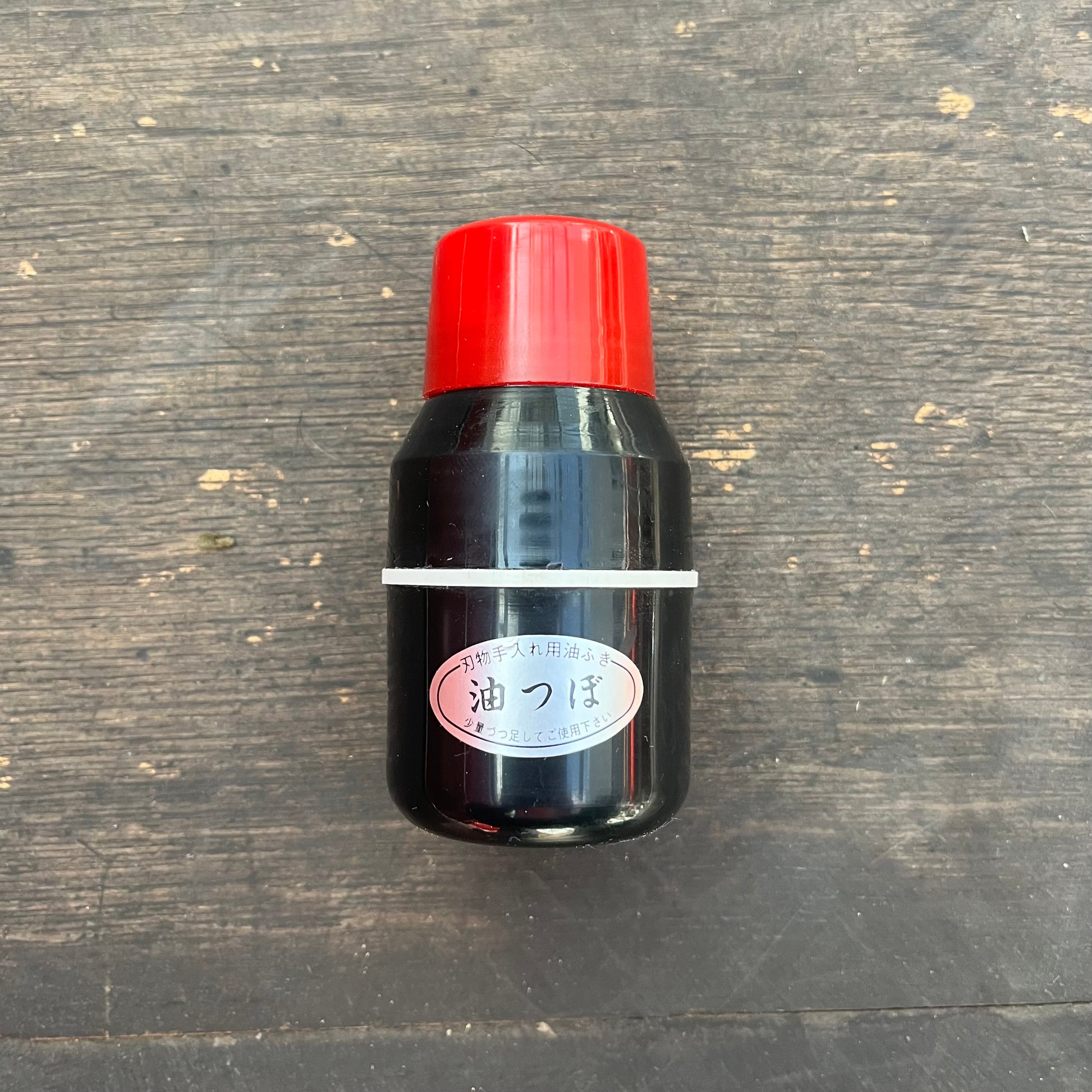 Ein kompakter schwarzer Niwaki | 'Oil Dispenser' Öl Spender ohne Inhalt von Niwaki, mit roter Kappe und weißem japanischen Etikett, sitzt auf einer zerkratzten dunklen Holzoberfläche - perfekt für präzise Pflanzenpflege oder Gartenpflege.