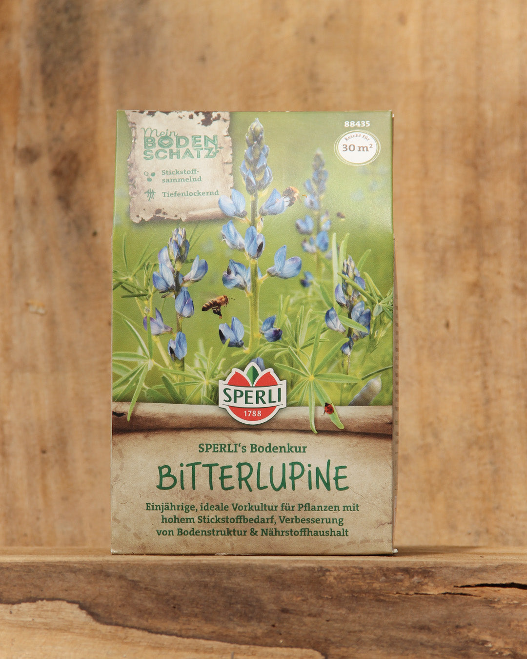 Eine 600 g-Packung Sperli's | Bitterlupine Bodenkur Saatgut steht auf einer Holzfläche mit blauen Lupinenblüten und Grünzeug. Der deutsche Text hebt die Wirksamkeit des Saatguts zur Bodenverbesserung hervor. Marke: Sperli's.
