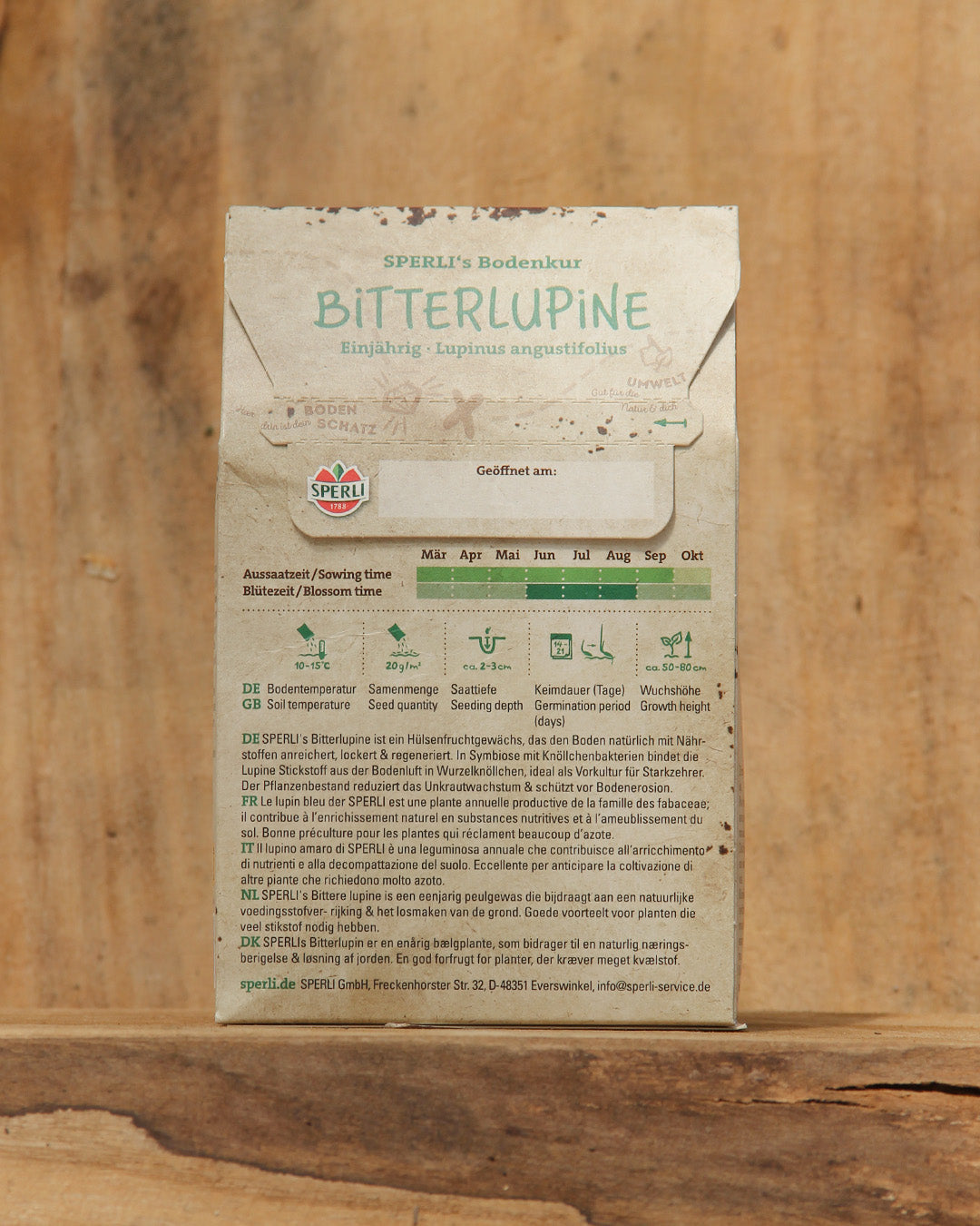 Eine 600-g-Packung Sperli's | Bitterlupine Bodenkur Saatgut steht aufrecht auf einem warm getönten Holz. Das Etikett enthält Piktogramme, Aussaatanweisungen in deutscher Sprache, Platz für das Öffnungsdatum und hebt die Verwendung für Gründüngung und Bodenverbesserung hervor.