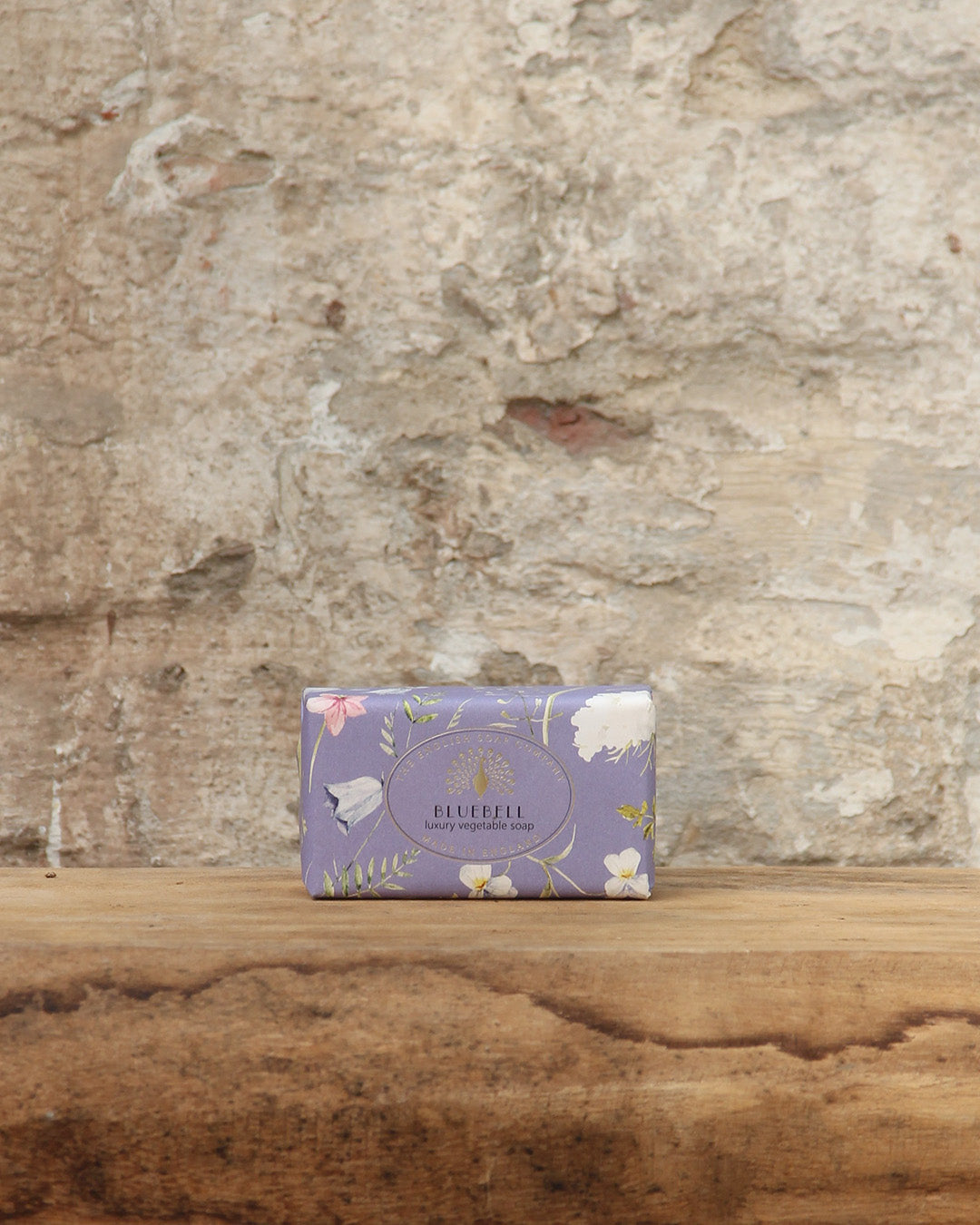 Ein lavendelfarbener Riegel ESC | 'Bluebell' pflanzliche Handseife mit Glockenblumenduft von The English Soap Company mit floralen Motiven ruht auf rustikalem Holz vor einer verwitterten Steinwand.