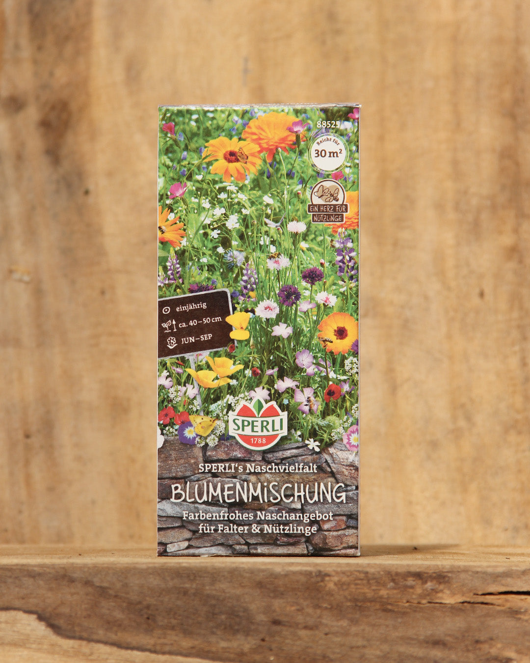 Eine farbenfrohe Saatgutpackung mit der Aufschrift Sperli's | Naschvielfalt Blumenmischung für Falter & Nützlinge zeigt blühende Wildblumen und Pflanzensymbole mit dem Markenzeichen von Sperli's auf einem Holzhintergrund.