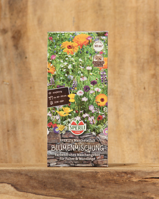 Eine farbenfrohe Saatgutpackung mit der Aufschrift Sperli's | Naschvielfalt Blumenmischung für Falter & Nützlinge zeigt blühende Wildblumen und Pflanzensymbole mit dem Markenzeichen von Sperli's auf einem Holzhintergrund.