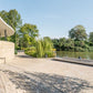 Ein modernes, geschwungenes Gebäude mit hellen Steinwänden blickt auf eine gepflasterte Terrasse an einem ruhigen Fluss, umgeben von Grün - eine inspirierende Szene, die in "Landschaftsarchitektur 2025" aus dem Callwey Verlag vorgestellt wird. Bänke säumen das Wasser; zwei Personen entspannen sich in der Nähe.