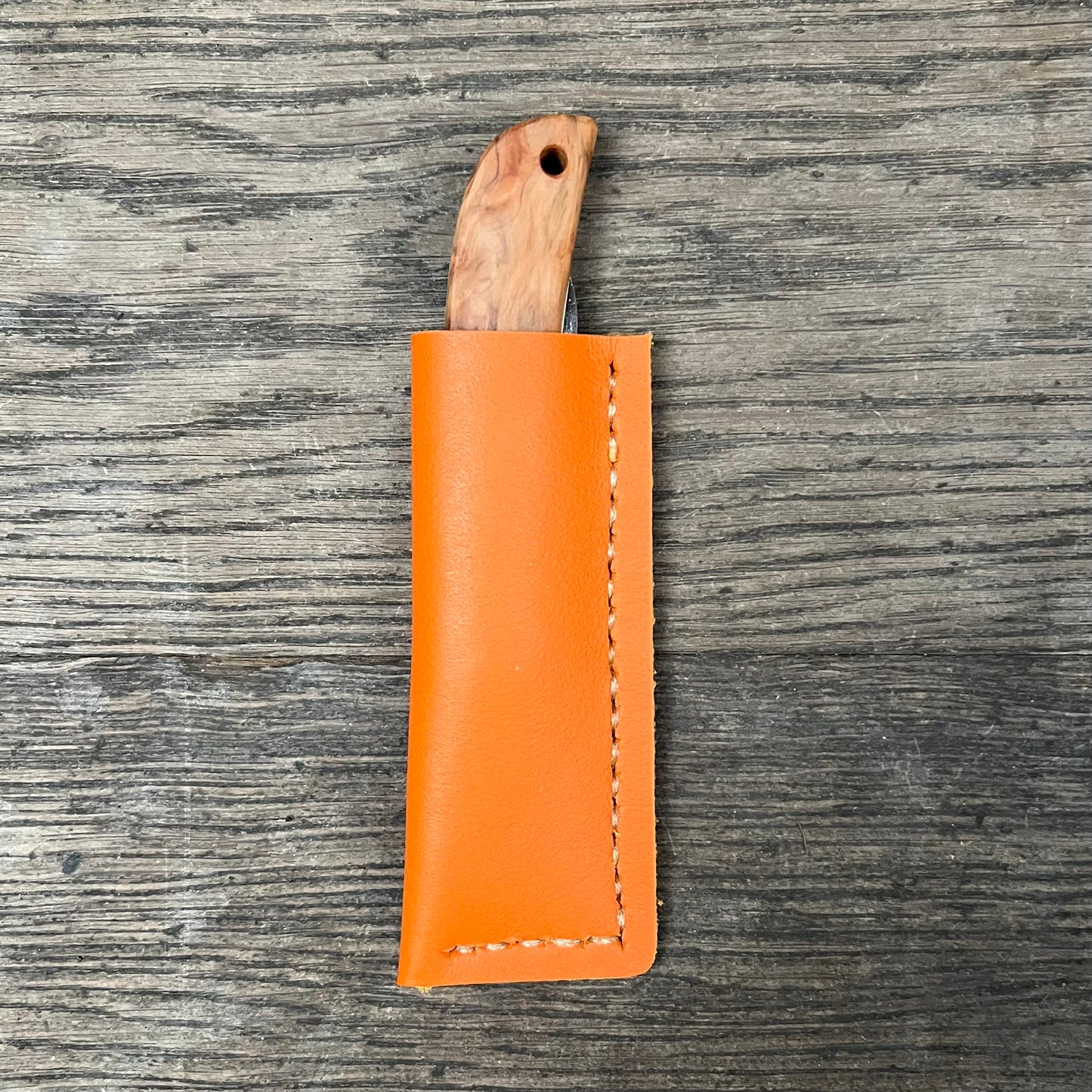 Das 'Kotoh X Niwaki Knife' Taschenmesser von Niwaki mit Holzgriff liegt teils in seiner orangefarbenen Lederschutzhülle auf Holz - ideal für Gartenarbeit oder als zuverlässiges Alltagsmesser.